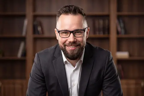 AI example headshot: smiling man with glasses and a beard in a dark suit in a law library